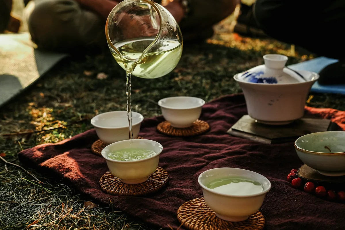 Thé vert versé d'une verseuse en verre dans de petites tasses blanches, sur un tapis en plein air